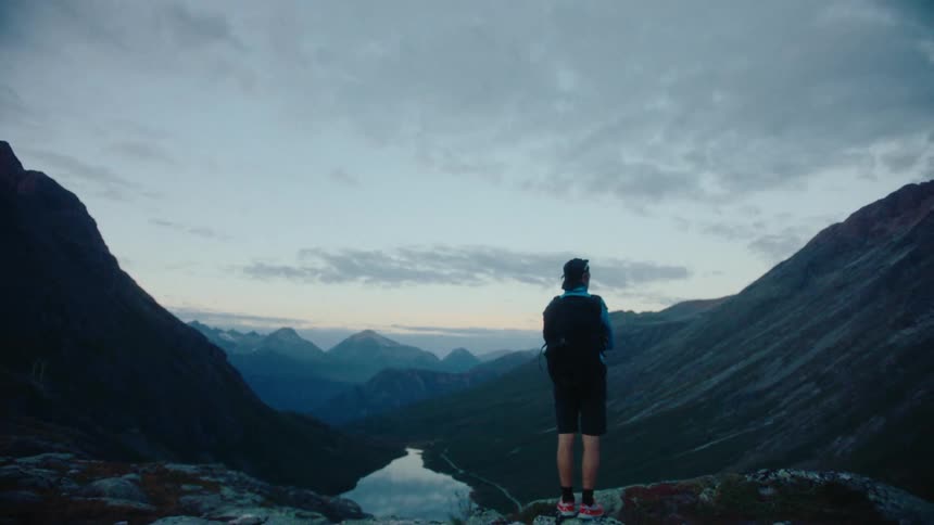 A man on top of a mountain peak observing the lake and the natural landscape with a mountain range