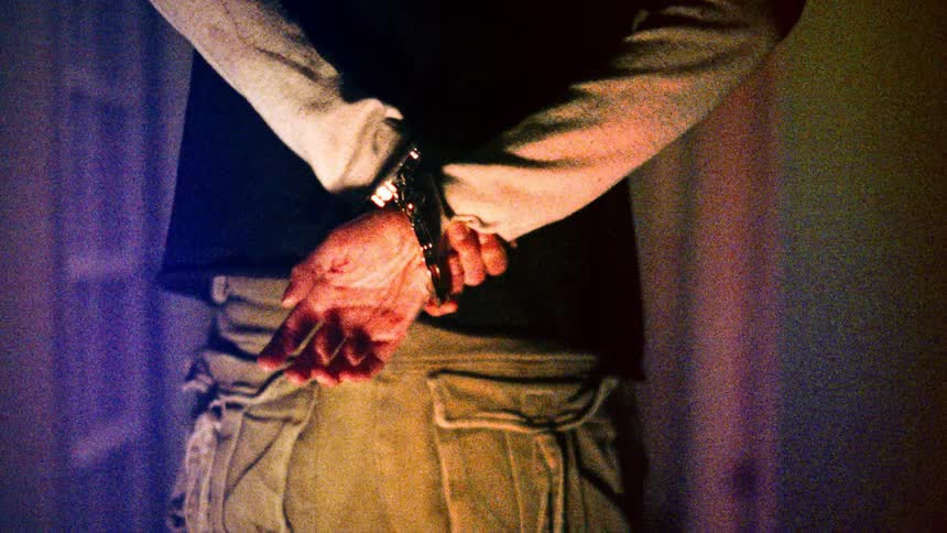 Handcuffed man with black shirt and green pants standing