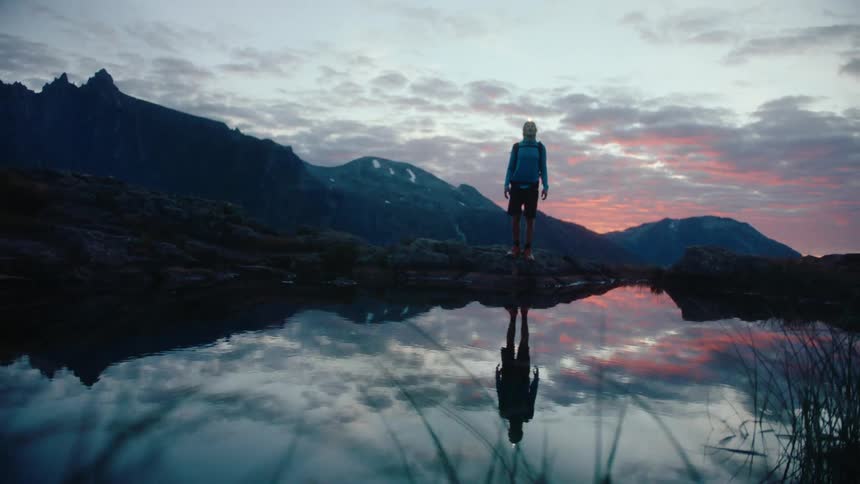 A man walking alongside a lake surrounded by a mountain range during a sunrise
