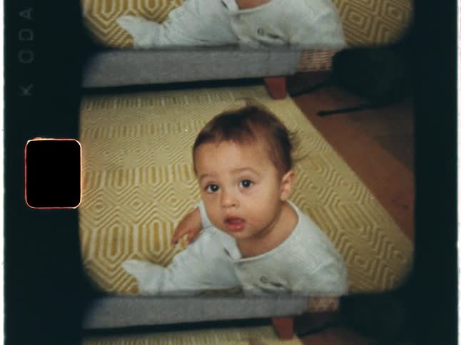 Super 8 film footage of a baby is sitting on the floor waving their hand