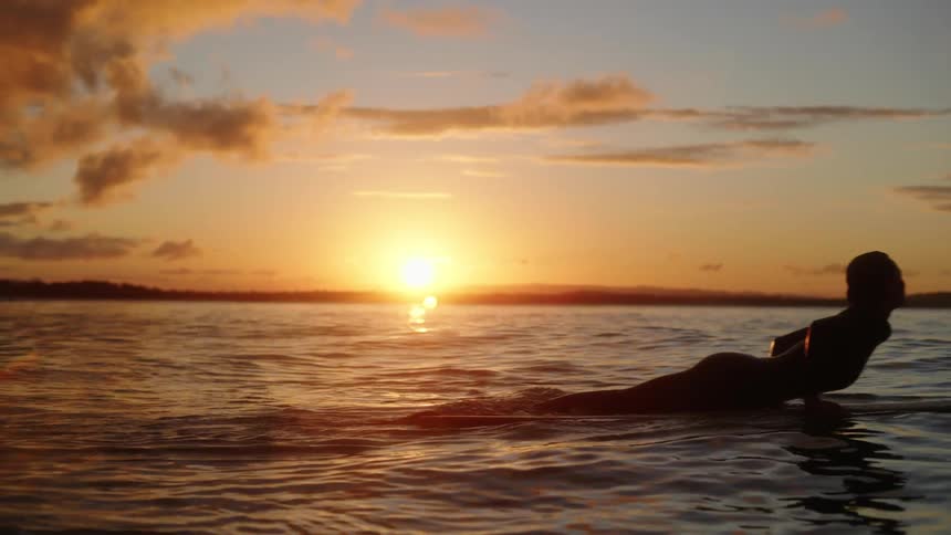 A woman swimming with her surfboard at sunset