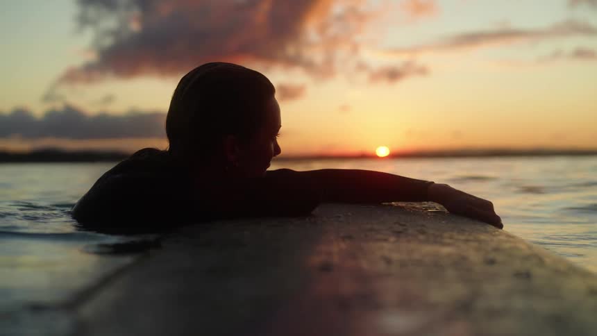 Woman leans half across surfboard while floating in ocean during sunset