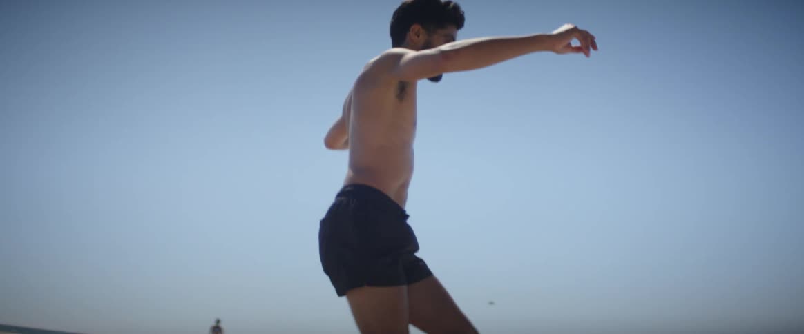 A shirtless man wearing skating shoe dancing near a beach at midday