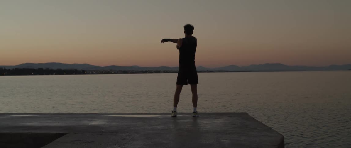 A man on a seaside dock with his back turned doing boxing moves and then stretching his arms during a sunrise