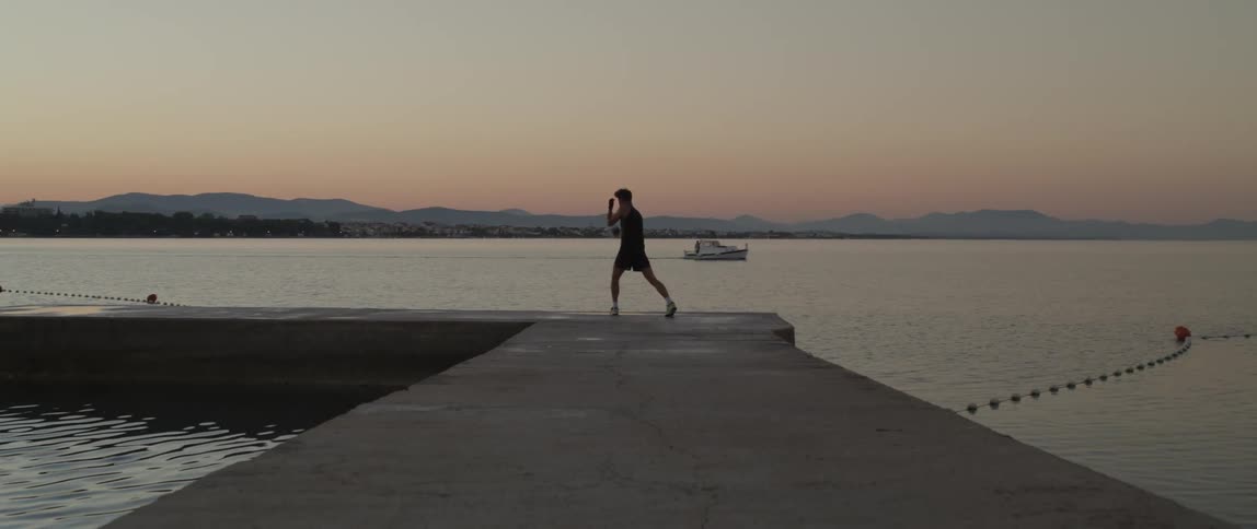 Male boxer runs along stone pier and shadowboxes as boat crosses under red tinted sky
