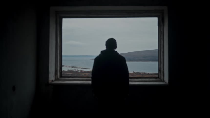 A man looking out the window at the sea and the coastline on a cloudy day