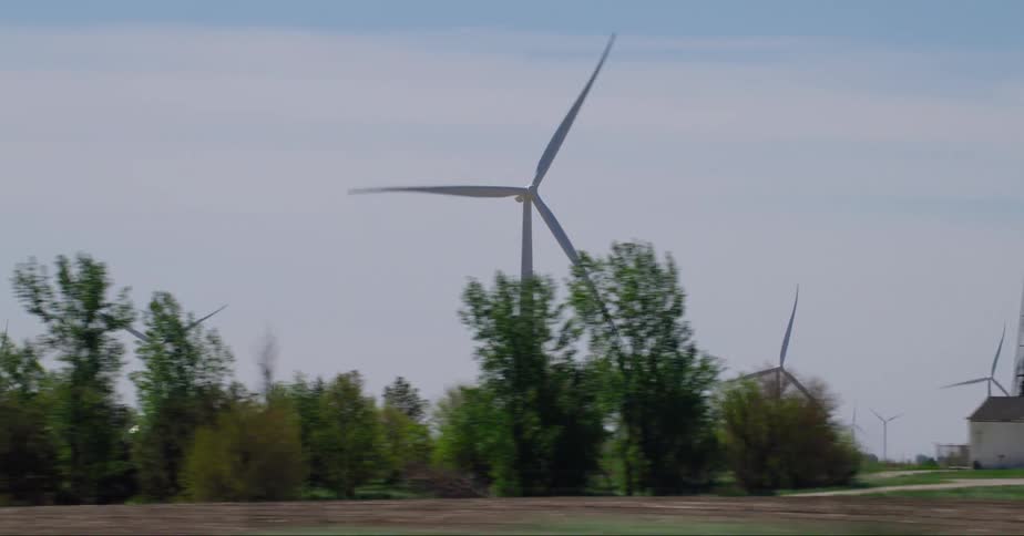 Wind turbines spin on a wind farm in a rual area among houses and trees and a utlity tower