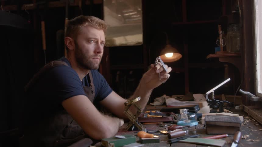 Clockmaker with beard and dark blue t shirt holds wristwatch and thinks