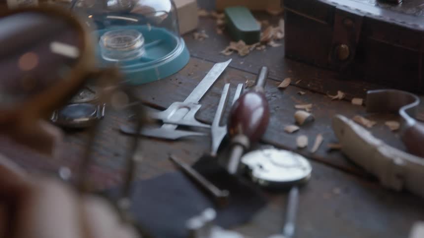 A mans hand fixes an object under a magnifying glass on a table with several scattered tools