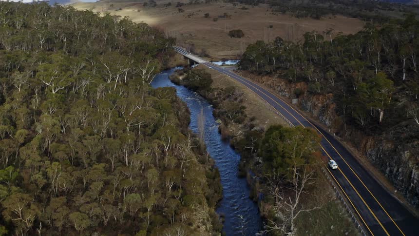 An aerial view of a vehicle driving on a road alongside a river and mountains