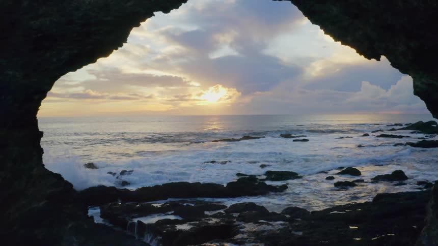 A sunrise or sunset is visible over the foamy white water of waves breaking on rocks as we move through a large rock archway that is nearly heartshaped to focus on the clouds sky sun