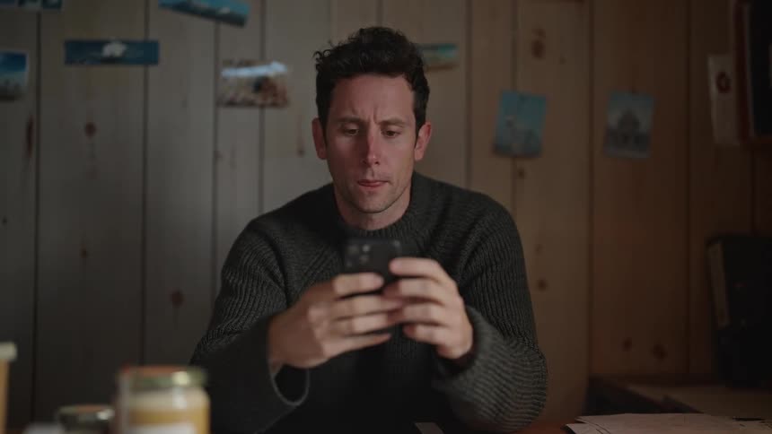 A man is sitting and placing his elbow on a dining table while operating his mobile device