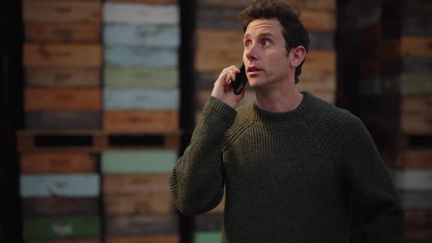A man stands in a warehouse in front of several stacked palettes while talking on a smartphone