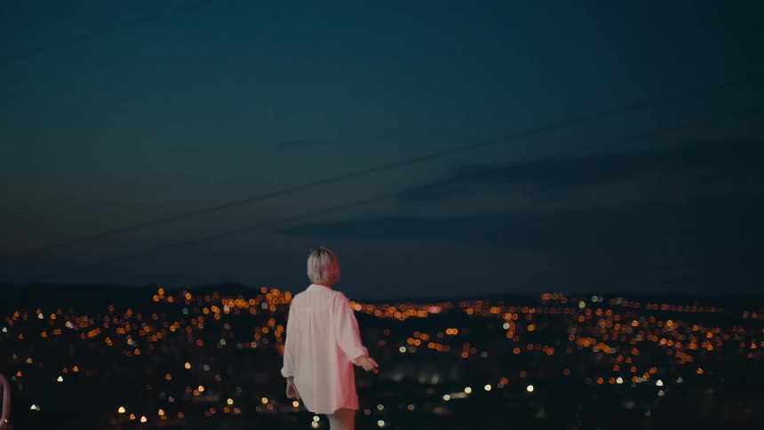 Blonde woman with white shirt and gray pants watches distant illuminated city scene