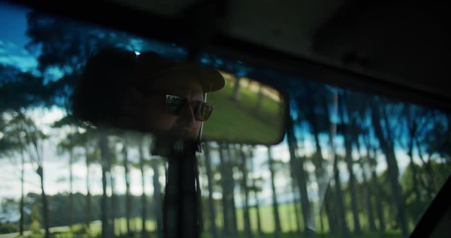 A mans head is shown in the rearview mirror while he drives past a heavily treed area