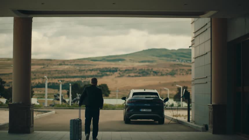 A man walks towards a car parked at a charger he is pulling along a suitcase on wheels unlocks the car as he approaches it there are mountains and a few trees and a road with traffic in the background