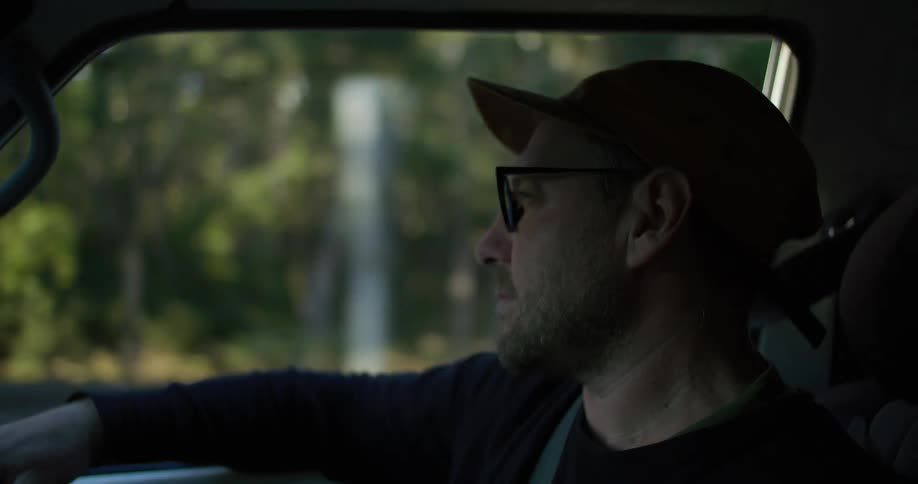 Man sits in passenger seat of car driving past woods