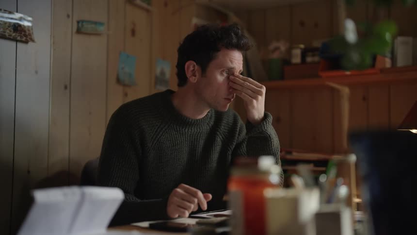 A man sitting indoors placing his hand on his face in a thoughtful expression