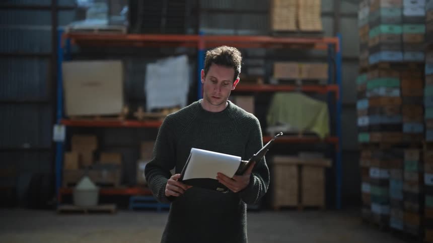 Man walks down warehouse aisle checking inventory against paperwork