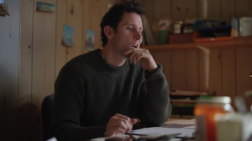 A man sits at a desk thinking before closing a folder and stretching