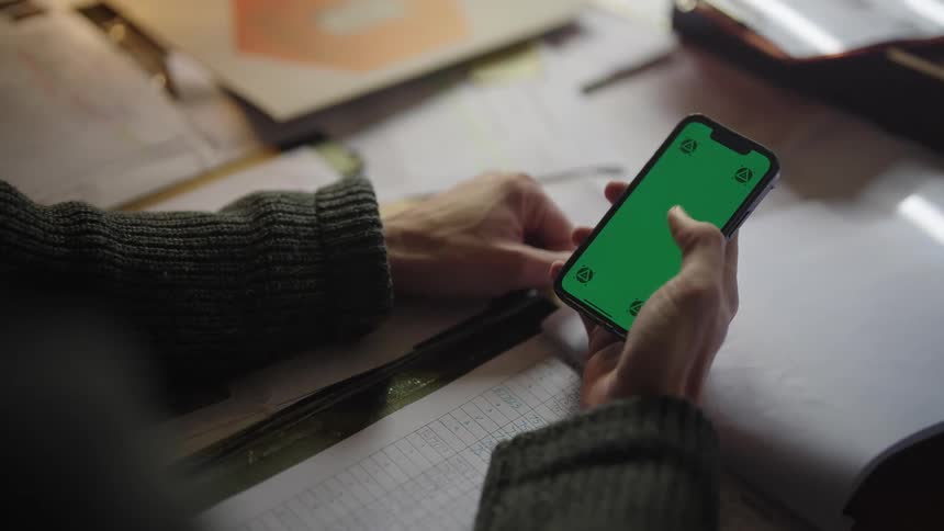 A man sitting at a desk holds a phone as he moves his thumb across the green screen