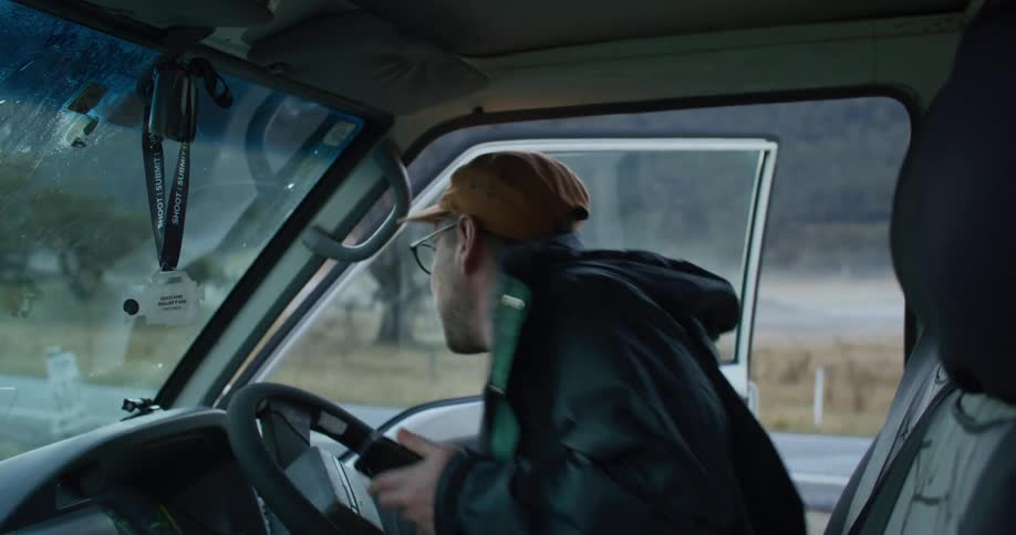 A man with a cap and reading glasses gets into a car and starts typing on his cell phone