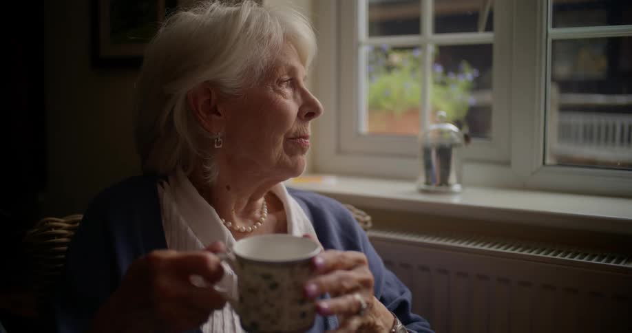 An elderly woman holding a mug is looking out the window