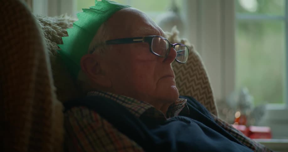 A man wearing a green paper Christmas crown sleeps in the chair