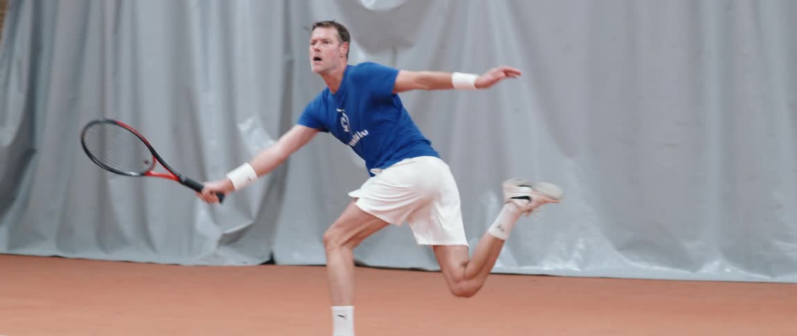 A man on a clay tennis court lunges in slow motion to hit a tennis ball