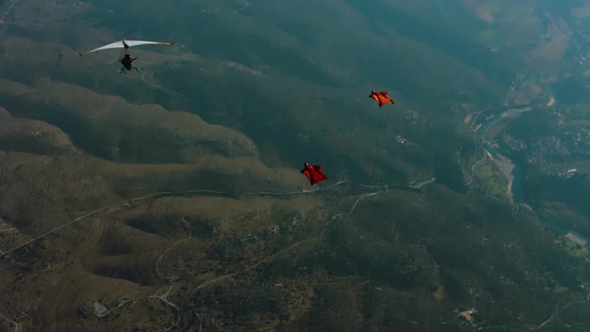 Two skydivers gliding in the sky without the parachute out yet flying over a vegetation area