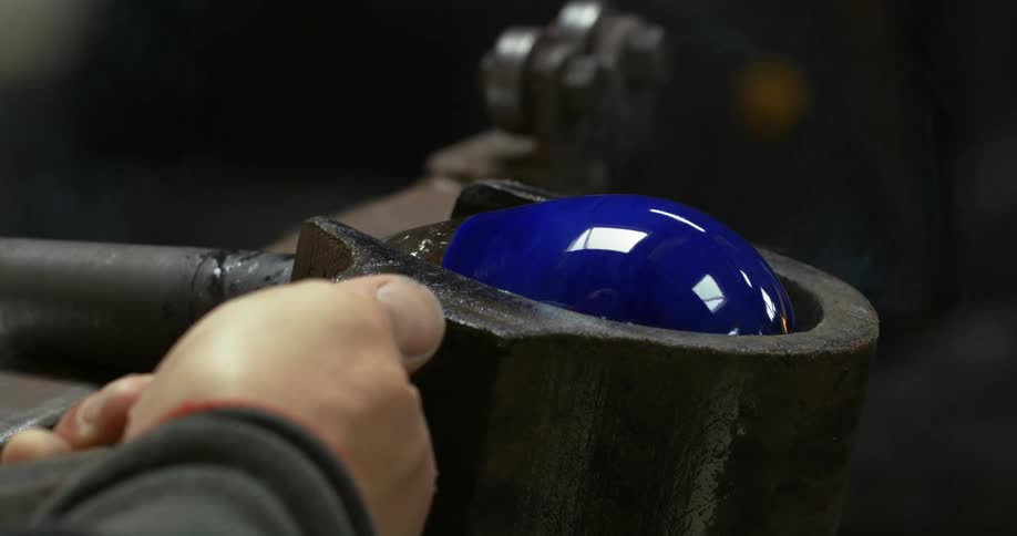 A hand guides a mold forming a globe of heated blue glass at the end of a metal pole being rolled down a thin metal beam
