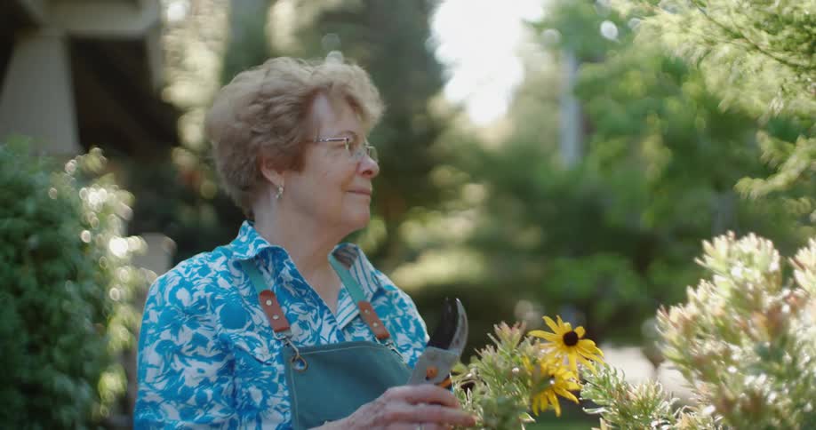 Woman with glasses earrings blue shirt and green apron tends to the plants in the backyard