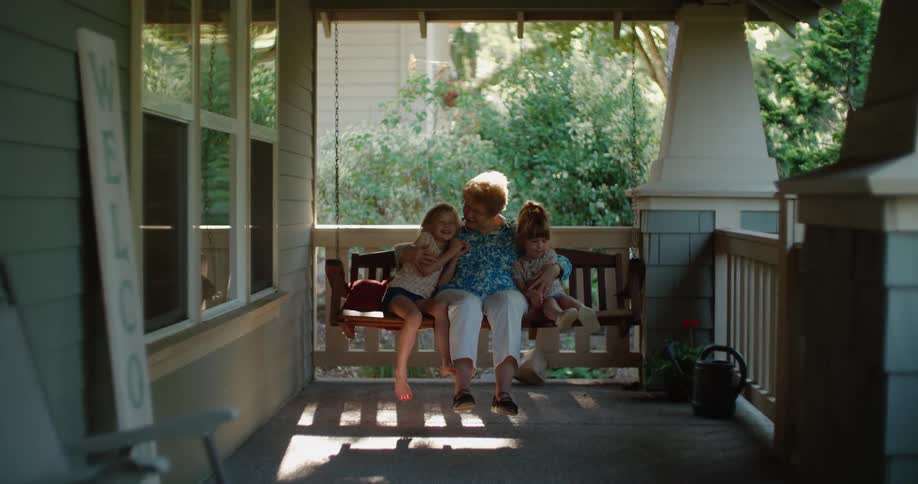 A grandmother and two granddaughters sit on a swinging bench on the porch