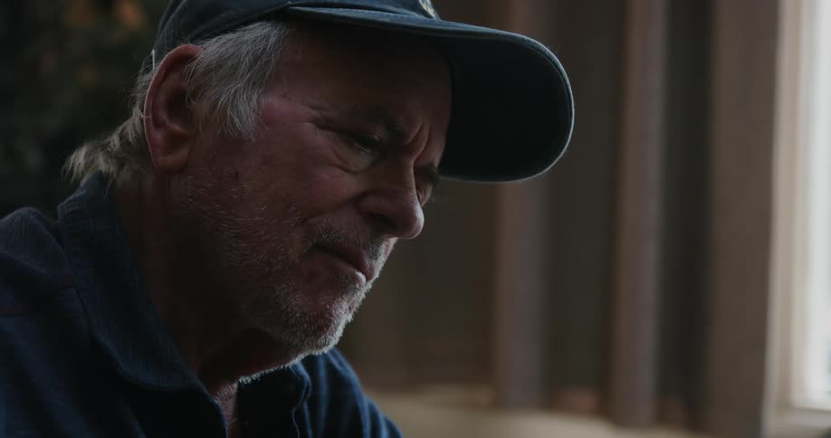 Man with gray hair a dark blue cap and a shirt sits at table thinking