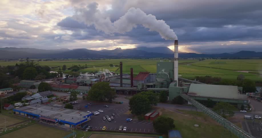 A tall smokestack overlooks a rural factory with cars in the parking lot and open fields with mountains in the distance under a cloudy sky