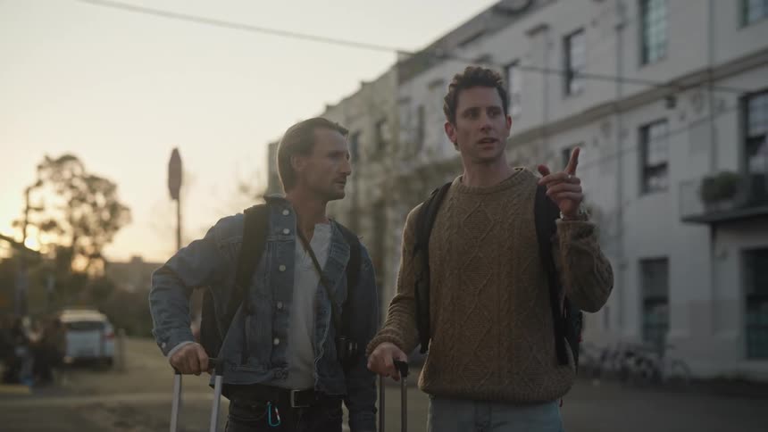 Two men are walking on a city sidewalk pulling their luggage and talking as they stop and one man pulls out his cell phone