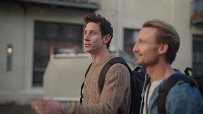 Two men with backpacks on their shoulders are walking and laughing and one of them puts their finger on the front