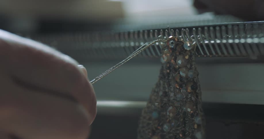 Two hands are using a metal contraption with a hook and some silver thread to make a gemstonefilled piece of lace
