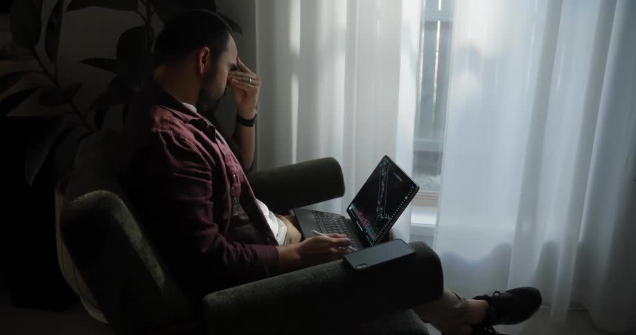 Tired man with a beard and a red shirt sits in armchair near the curtains in a room