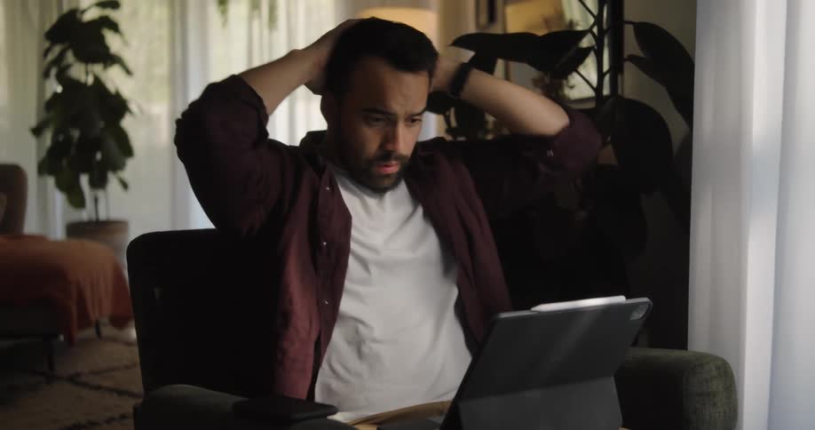 A man is sitting in a living room chair typing on a laptop computer and he stops to think as he leans back and puts his hands behind his head
