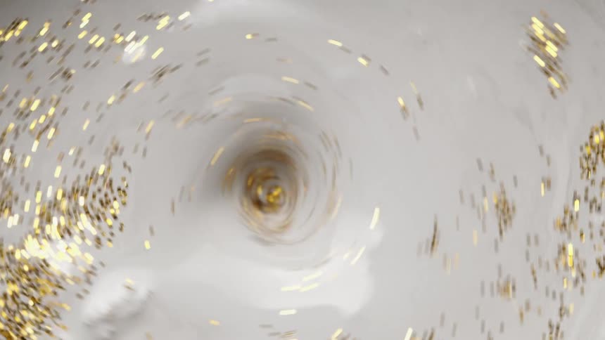 Golden glitter floats on the surface of and into small whirlpool of a pale fluid