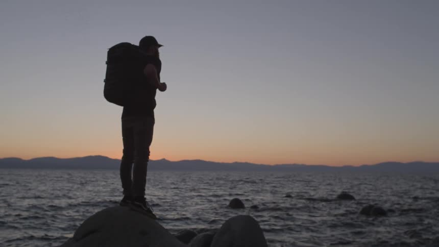A man is perched on a rock in the water at sunset