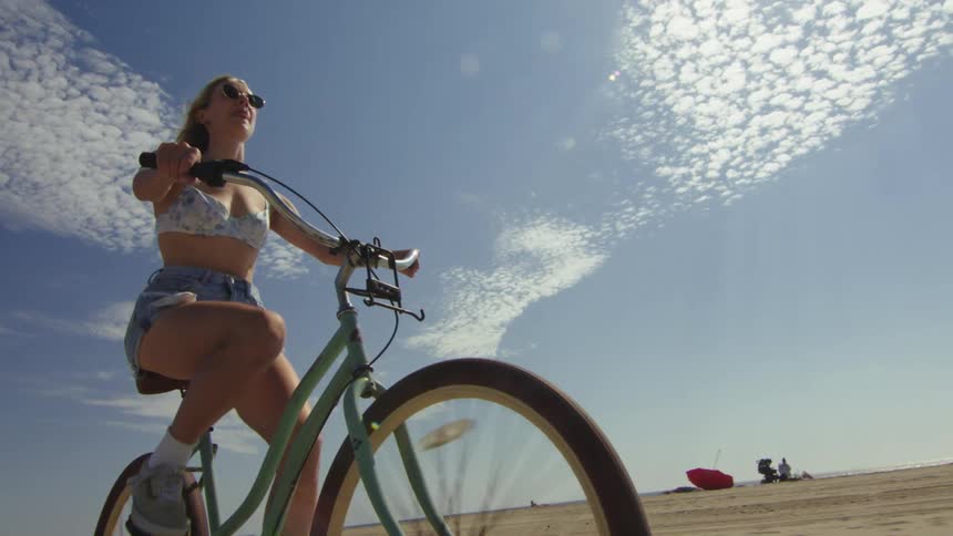 A smiling woman in shorts and a short top is riding a bicycle near a beach