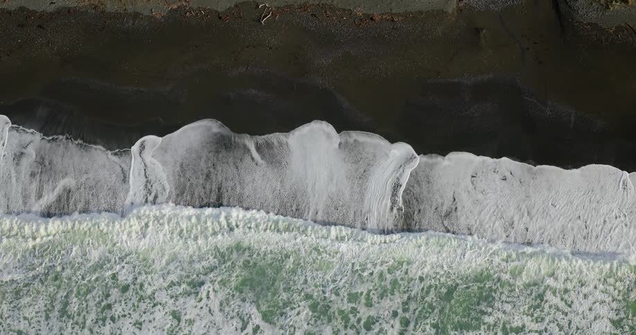 Aerial view of the sea with moving waves touching the black areas of the beach