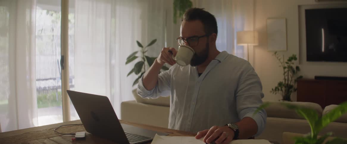 Man drinking coffee from a mug sitting in front of a laptop at a table in his living room