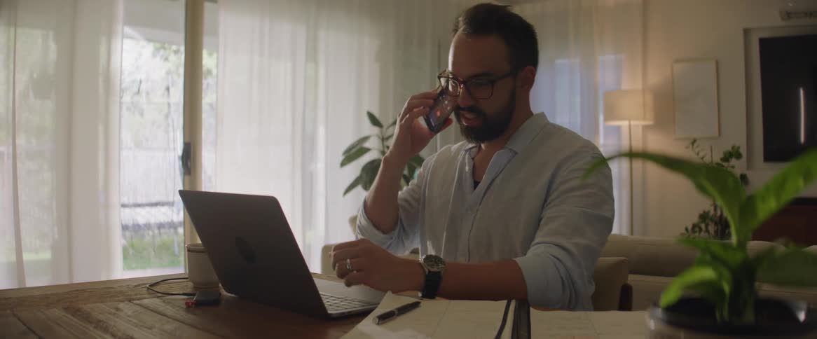 Man sitting at a table in front of his laptop before picking up his cellphone and talking on it while smiling