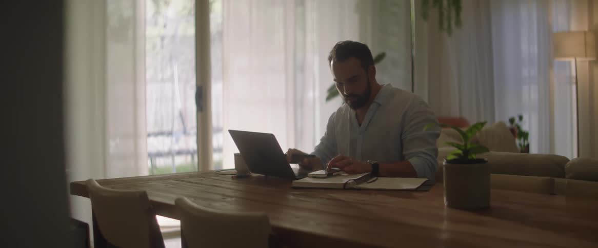 A man at a desk is typing on the computer and taking notes