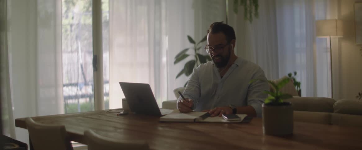 Man sitting at his dining room table in front of his laptop while nodding at the screen and writing on a note pad