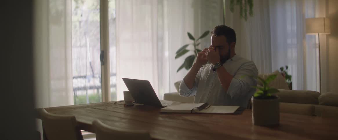 Man sitting at a long wooden table talking on a cellphone in front of a laptop looking stressed