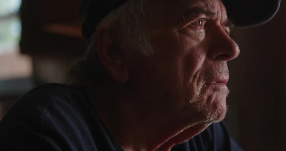An elderly mans face as he looks contemplatively furrowing his brow and wiping his face
